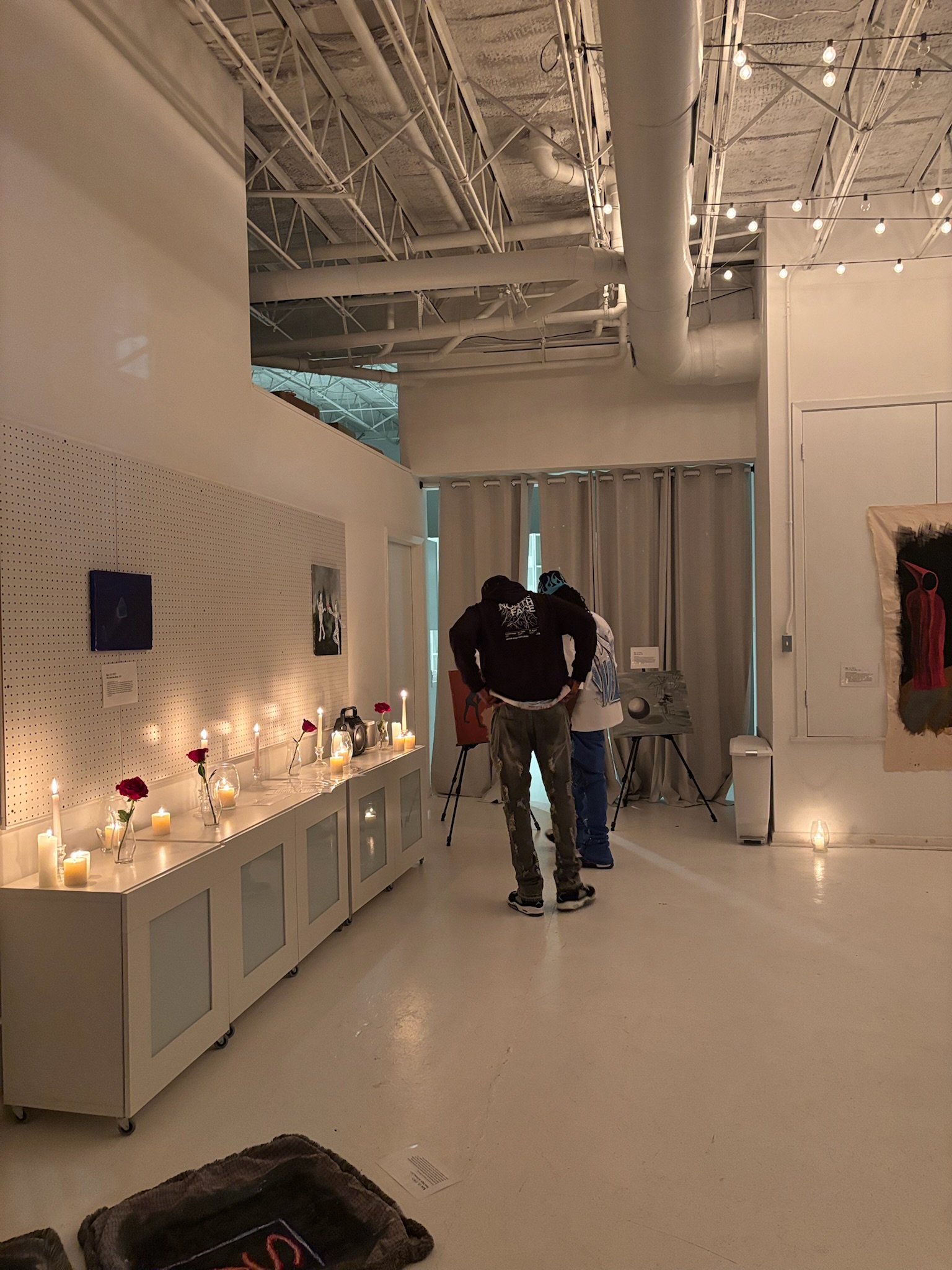 Installation view showing candles lining a white cabinet with artworks on easels in the background